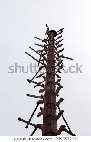 A vertical, low-angle shot of a tall, rustic wooden totem or memorial pole adorned with numerous antique mining pickaxes and axes pointing outward at various angles. The weather-beaten wooden post stands tall against a flat, white overcast sky, with the dark green tips of pine trees visible at the base of the frame. This poignant structural monument serves as a historical tribute, with each individual pickaxe representing a miner who tragically lost their life working underground in the local iron mines.