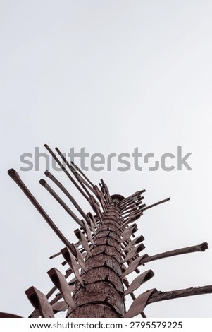 A vertical, low-angle shot of a tall, rustic wooden totem or memorial pole adorned with numerous antique mining pickaxes and axes pointing outward at various angles. The weather-beaten wooden post stands tall against a flat, white overcast sky, with the dark green tips of pine trees visible at the base of the frame. This poignant structural monument serves as a historical tribute, with each individual pickaxe representing a miner who tragically lost their life working underground in the local iron mines.