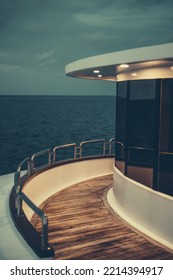 A Vertical Low-key View Of A Diving Safari Yacht's Middle Deck With A Chrome Fence, Lit Wooden Floor, Illuminated Ceiling, And Windows Reflecting The Dark Ocean Waterscape And Part Of The Evening Sky