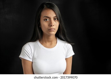 vertical medium close-up of a serious and beautiful black haired Latina woman wearing a white t-shirt on a black background with copy space.