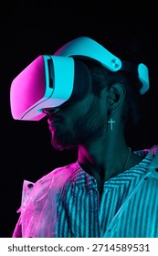Vertical medium close-up studio portrait of stylish young Indian man wearing VR headset standing in neon light in darkness