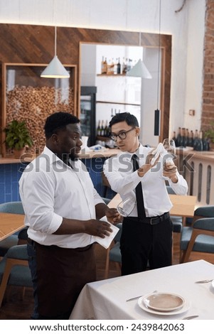 Vertical medium long shot of young African American and Asian waiters talking while polishing glass wines