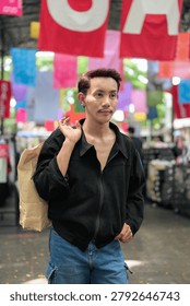 A vertical medium portrait shot centered on a non-binary gay man of Isan, Thai descent standing in an outdoor market setting. The model has short dark hair with reddish highlights, a mustache, black stud earrings on both ears, and is dressed in a casual black jacket paired with blue denim cargo shorts. The individual is looking away off-camera to their right with a calm expression while holding a large, crumpled brown paper shopping bag over their right shoulder. The background features colorful fabric banners and signs hanging overhead, rendered out of focus. Shot with soft, natural outdoor daylight that highlights the model's authentic, unretouched skin features and personal style. The composition is vertically balanced with a shallow depth of field, keeping the subject in sharp focus.