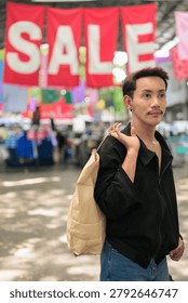 A vertical medium portrait shot of a non-binary gay man of Isan, Thai descent standing outdoors in a street market corridor. The model has short dark hair with subtle reddish-maroon highlights, a mustache, black stud earrings, and is wearing a black casual jacket. Positioned on the right side of the frame, the individual is looking away off-camera to their right with a serene facial expression while carrying a large brown paper shopping bag held by its handles over their shoulder. The background features huge, vibrant red fabric banners spelling out 