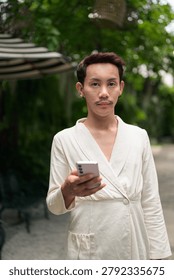 A vertical medium portrait of a stylish non-binary gay Thai man with styled red-brown hair and a mustache, dressed in a fashionable cream wrap cardigan jacket. He is standing outdoors in a lush garden environment, holding a pink smartphone in his hand while looking directly into the camera lens with a calm, neutral expression. The background displays a soft, shallow depth of field blur featuring vibrant green tree leaves and a striped patio umbrella. This authentic, unaltered image captures contemporary LGBTQ+, queer, and gender-fluid representation, modern digital connectivity, and casual daytime lifestyle in Southeast Asia.