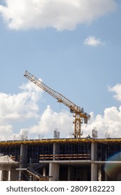 A vertical, medium shot capturing an active building construction site in Astana, Kazakhstan. A prominent yellow tower crane stands tall atop a monolithic concrete structure under development, with reinforcement bars visible. Construction workers in helmets are working on the upper level next to scaffolding. The scene is shot on a bright summer day against a blue sky filled with fluffy white cumulus clouds.