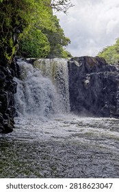 A vertical, medium shot capturing the natural force of the Grand River South East (GRSE) waterfall in Mauritius. The water cascades heavily over a jagged, dark volcanic basalt cliff face into a churning river plunge pool below