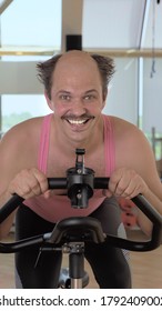 Vertical, Medium Shot, Funny Bald Man Is Training On An Exercise Bike, Smiling.