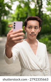 A vertical medium shot of a stylish non-binary gay Thai man and influencer with styled red-brown hair and a mustache, dressed in a fashionable cream wrap cardigan jacket. He is standing outdoors on a garden pathway, holding up a pink smartphone to take a selfie or record a video call while looking away thoughtfully to the side. The green park background features a soft, shallow depth of field bokeh with lush trees and a distant decorative circular light. This authentic, unaltered image captures contemporary LGBTQ+, queer, and gender-fluid representation, digital content creation, modern communication, and casual lifestyle in Southeast Asia.