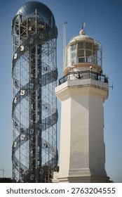 A vertical medium shot of two distinct structures standing side-by-side. On the left is the tall, modern Alphabetic Tower, a steel lattice structure wrapped in decorative alphabet characters, topped with a large, faceted glass sphere. On the right is a traditional, octagonal white lighthouse with a glass-paneled lantern room and a wrought-iron gallery. The two buildings are set against a solid, deep blue sky.