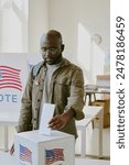 Vertical medium shot of young African American male citizen putting ballot paper into ballot box at polling place on election day