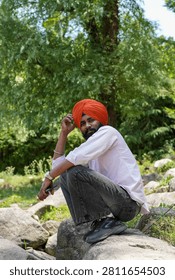 A vertical, medium shot of a young Indian Sikh man wearing a vibrant orange turban and a light-colored shirt, crouching on a large rock while looking at the camera amidst the lush greenery of the forests in Himachal Pradesh.