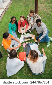 Vertical. Multiracial students using a laptop computert sitting in a circle on the grass, doing homework outside at university campus. Young high school people working on a academy task or schoolwork