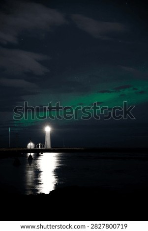 Vertical night composition capturing green aurora borealis shining above Grotta lighthouse with bright light reflecting on calm coastal water in Iceland.
