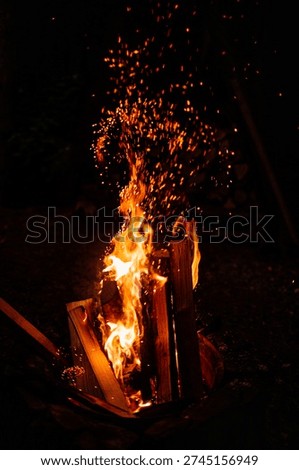 A vertical night shot capturing a vibrant campfire with intense orange flames and a massive shower of glowing golden sparks rising high against a dark, pitch-black background.