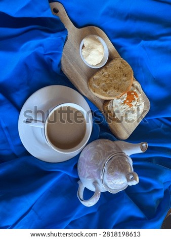 Vertical overhead flat lay of a cup of creamy coffee served beside an elegant floral ceramic teapot on vibrant royal blue fabric. Minimal beverage still life suitable for café, hospitality, and breakfast concepts with generous copy space.