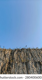 A vertical perspective looking up at a rocky retaining wall under a bright blue sky.