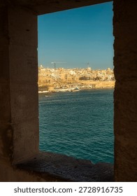 A vertical perspective looking through a thick limestone window frame at the historic city of Valletta. The scene features the deep blue Mediterranean Sea, sunlit historic buildings, and a clear blue sky, viewed from within an ancient fortification. Malta, Valetta, Isla