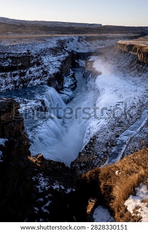 Vertical perspective showing the deep canyon cut by the frozen Gullfoss waterfall in Iceland. Snow-covered rocky cliffs frame the rushing turquoise water and rising mist.