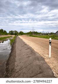 Vertical perspective view of a newly constructed rural gravel road on a high earth embankment. Detailed outdoor shot of fresh road infrastructure with a smoothed soil slope, a drainage ditch with water, and a white roadside delineator post under a dramatic cloudy sky.