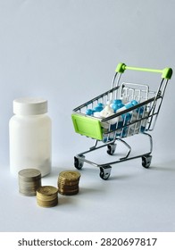 Vertical perspective of a white pill container, stacked money coins, and a miniature shopping cart filled with medical capsules.