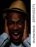 Vertical photo of an African-American man in a straw hat and a blue shirt with a mad face from pain, he holds his head with his hands with his mouth open from shock on a black background