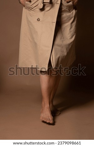 Vertical photo. Bare female feet. White tanned woman in a beige raincoat on a brown background in the studio. Concept of quiet walking, tiptoe, silence, beauty, shoe shopping, naturalness, epilation
