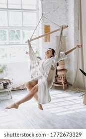 Vertical Photo Of Carefree Young Adult Female In Bathrobe Sitting On A Hanging Swing At Home, Enjoying Lazy Weekend Morning, Having Fun And Raised Hands Up, Feeling Free