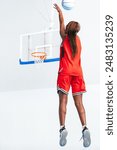 Vertical photo a female basketball player in a red uniform executes a jump shot towards the hoop on an indoor court, demonstrating her athleticism, precision, and skill during a practice session.