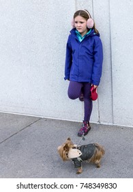 Vertical Photo Of Forlorn-looking Little Girl In Fall Jacket And Ear Muffs Leaning With One Foot On Wall While Holding Tiny Yorkshire Terrier Dog On Leash