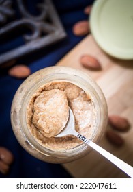 Vertical Photo Of Homemade Almond Butter On A Spoon Placed In A Glass Jar. Jar Stands On A Wooden Cutting Board Placed On A Dark Blue Table Cloth. 