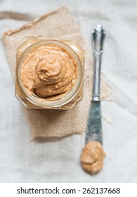 Vertical photo of homemade natural almond butter in a glass jar placed on rustic beige table cloth and with a butter knife on the side. Pastel colors. View from above. 