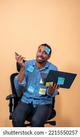 A Vertical Photo Of A Latino Man With A Sorry Face Holding A Phone And A Laptop. Wearing A Blue Shirt And Sitting In An Office Chair. A Lot Of Sticky Notes Glued On Him.