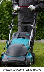 Vertical Photo Of A Man In Overalls And White Gloves Mowing Green Grass In The Garden With A Lawn Mower. A Worker In Coveralls Uses A Modern Lawnmower In The Garden. Professional Lawn Care Service.