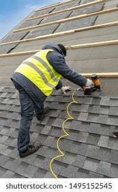 Vertical Photo Of Professional Collar Worker Or Roofer In Special Protective Work Wear Using Air Or Pneumatic Nail Gun And Installing Asphalt Or Bitumen Shingle On Top Of The Roof Under Construction 