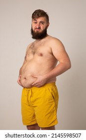 Vertical Photo With A Sad Fat Man. An Overweight Young Man In Yellow Shorts On A Gray Background Dejectedly Holds And Feels His Stomach