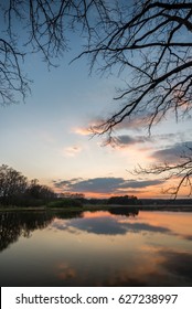 Vertical photo with spring evening landscape after sunset. Cloudy sky is nicely reflected in the water of small pond. Grass, reed, threes and branches are in background and foreground.