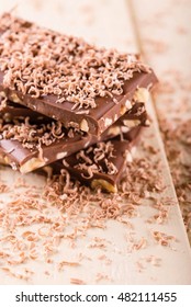 Vertical photo of stack from brown chocolate which contains nuts. Sweet chocolate shavings are spilled everywhere on the picture. Dessert is placed on wooden board with light color.