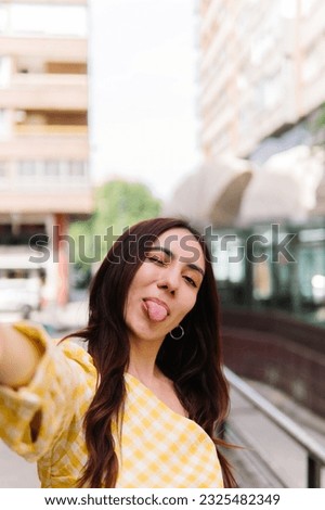 Vertical photo of a stylish girl shows her tongue while taking a selfie in the street