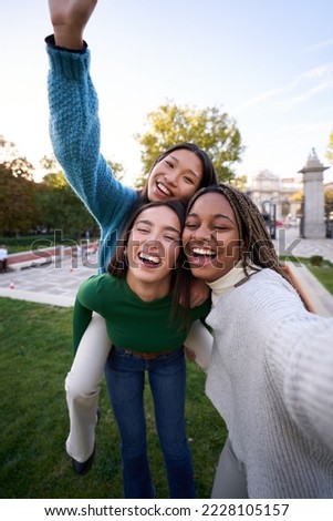 Vertical photo of three multicultural female friends taking smiley selfie doing piggyback. Young women having fun together outdoors at city park in Madrid. People enjoying travel in weekend holidays.