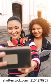 Vertical Photo Of Two Multiracial Latina Students Taking A Self-portrait With Their Cell Phone Smiling Outdoors In A Public Square In Leon