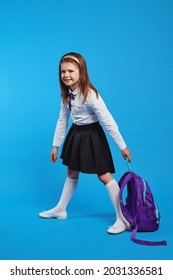 Vertical photo of unhappy schoolgirl in uniform holding a heavy backpack, isolated against blue background. Problem of weighting student accessories