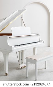 Vertical Photo Of White Grand Piano With Opened Lid And A Piano Bench In A Small White Studio At Music School