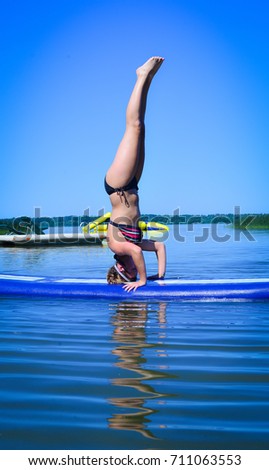 Vertical photo of young woman doing hand stand on stand up paddle board. She is wearing a bikini on a sunny day blue lake and sky. 
