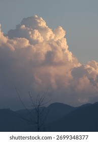 vertical photograph capturing a dramatic, large cumulus cloud formation that is brightly lit by the golden light of the setting or rising sun, towering above a ridge of layered, shadowed mountains wit