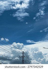 Vertical photograph of dramatic white clouds and deep blue sky above power lines and utility poles. Atmospheric outdoor scene capturing weather, urban infrastructure, and natural cloud formations.