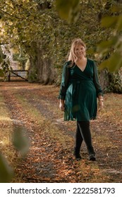 Vertical picture of a curvy woman wearing a dark green dress and black ankle boots; the woman is smiling walking along an alley covered with autumn leaves - Shutterstock ID 2222581793