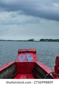 Vertical Point of View Shot from Bow of Rustic Red Wooden Boat Moving Across Vast Calm Waters of Dumbur Lake in Tripura India. An immersive vertical point-of-view (POV) shot taken from the front of a moving wooden boat on Dumbur Lake in Tripura, Northeast India. The foreground highlights the weathered red painted prow of the boat, featuring textured peeling paint that adds rustic charm. The boat cuts through the calm, rippling water, heading towards a distant tree-lined shoreline under a dramatic grey overcast sky. This image perfectly captures the experience of rural water tourism and exploration.