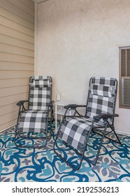 Vertical Porch Of A House With Two Lounge Chairs And Carpet With Blue And White Colors. There Is A Plant Beside The Watering Can On The Left Near The Railings.