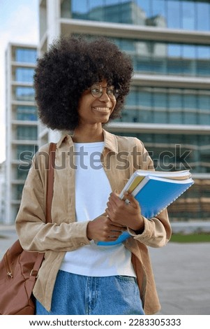 A vertical portrait of African American girl with curly afro hairstyle stands with backpack and holds exercise books in university campus. Smart student looking aside on city background, copy space.